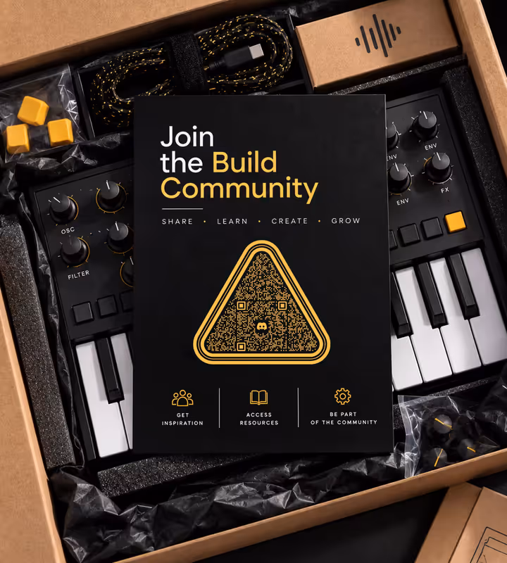 A black “Join the Build Community” card with a gold triangular QR code, lying on a synthesizer in an open kit box beside a braided cable and spare keycaps