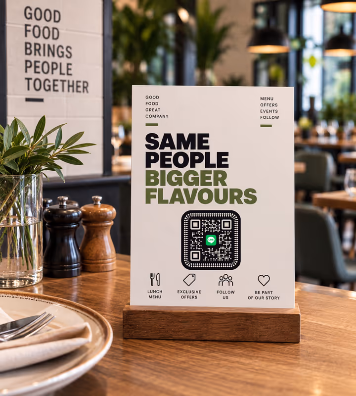 A white “Same people, bigger flavours” table card with a black rounded QR code on a wooden base, standing on a restaurant table
