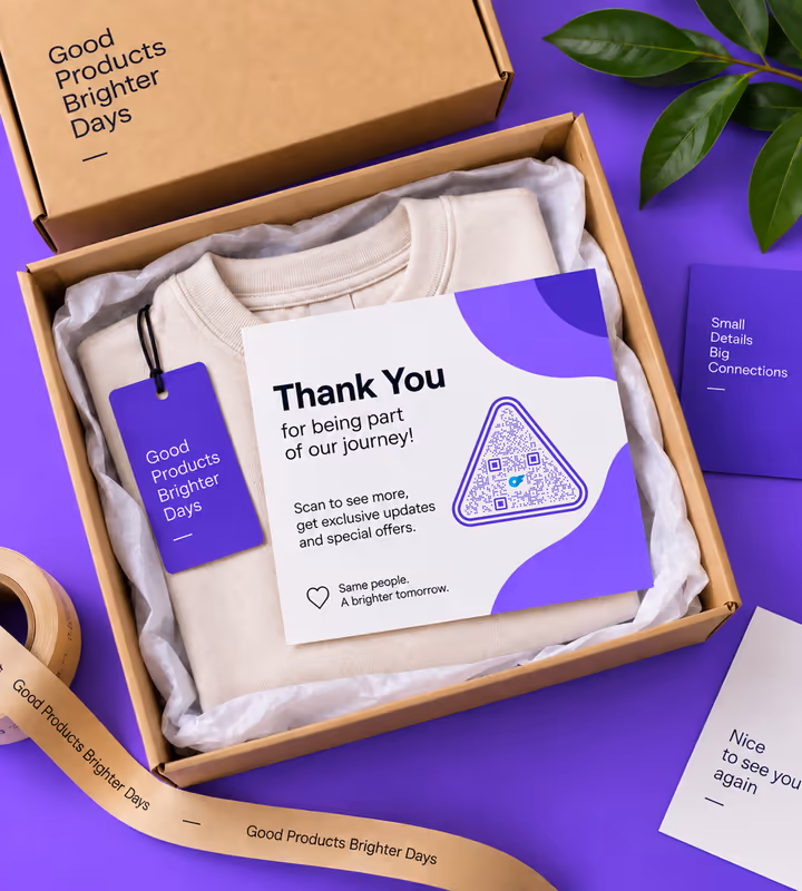 A purple and white “Thank you for being part of our journey” card with a triangular QR code, sitting in a shipping box on a folded sweatshirt