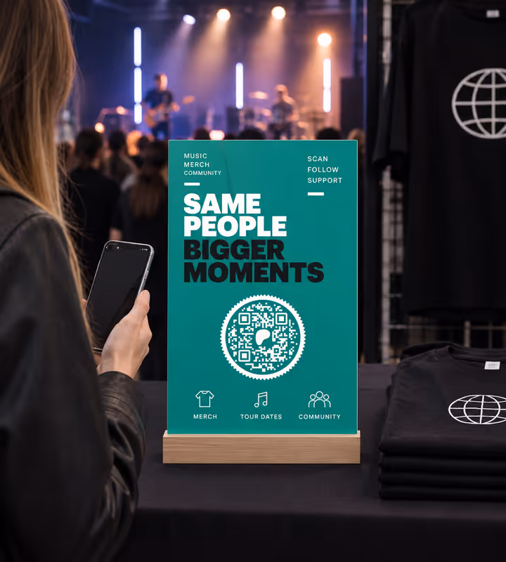 A teal merch-table sign reading “Same people, bigger moments” with a circular QR code, folded band T-shirts beside it and a gig in progress behind