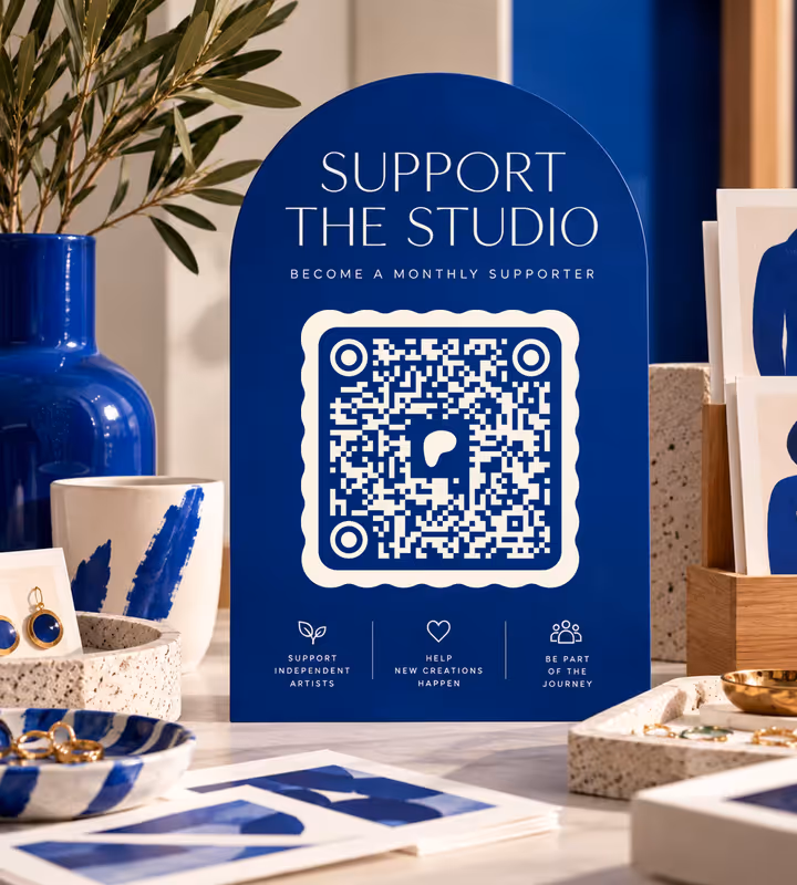 A blue arched “Support the Studio” sign with a cream wavy-edged QR code, on a studio table among blue-painted ceramics, prints and earrings