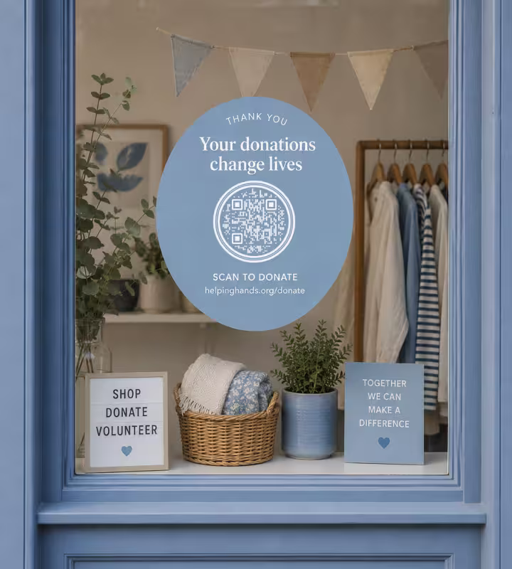 A round blue decal in a charity shop window reading “Thank you - your donations change lives” with a QR code marked “Scan to donate”, with bunting, a rail of clothes and a “Shop, donate, volunteer” sign inside