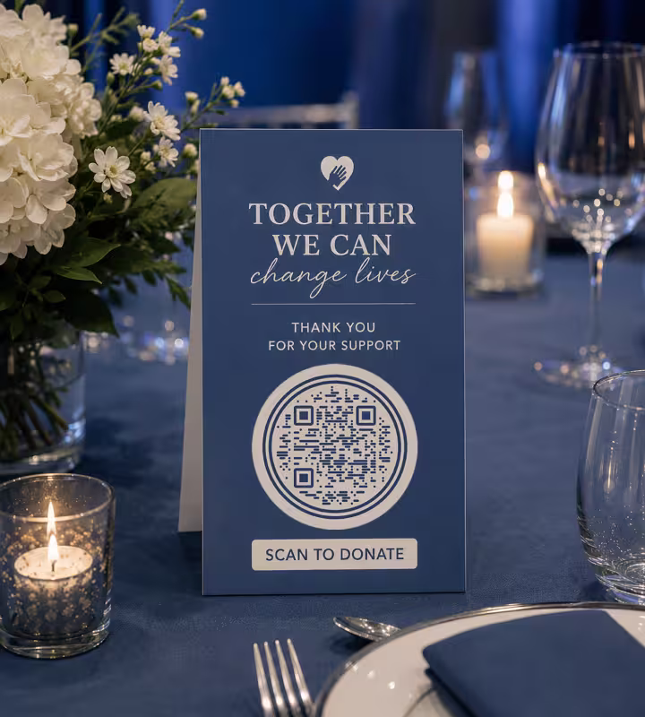 A navy table card at a fundraising dinner reading “Together we can change lives - thank you for your support”, with a round QR code and “Scan to donate”, among candles and glassware