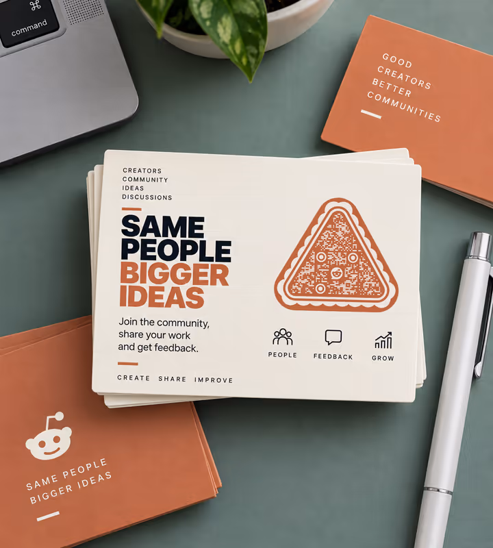 A stack of cream cards reading “Same people, bigger ideas” with an orange triangular QR code, on a green desk beside a laptop