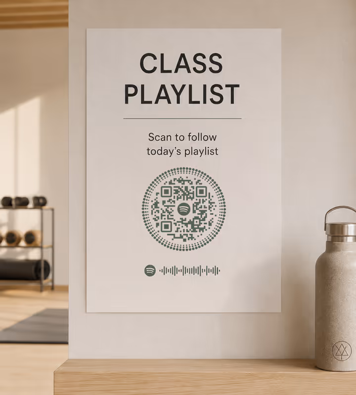 A studio wall sign reading “Class playlist - scan to follow today's playlist” with a circular dotted QR code, gym equipment behind