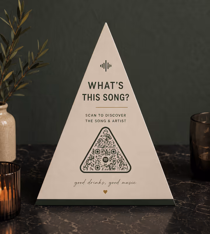 A triangular table sign asking “What's this song?” with a rounded triangular QR code, on a dark marble bar table beside a candle