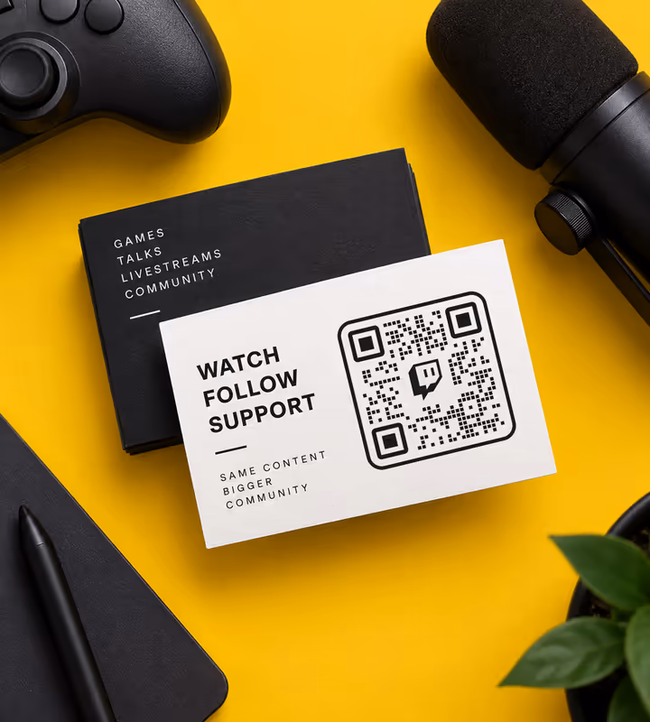 A white card reading “Watch, follow, support” with a square QR code, on a yellow desk beside a controller and a microphone