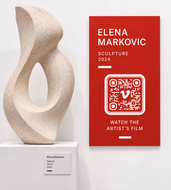 A red gallery wall label for “Elena Markovic, Sculpture 2024” with a white rounded QR code and “Watch the artist's film”, beside a stone sculpture on a plinth