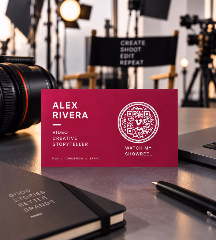 A magenta business card for “Alex Rivera, video creative storyteller” with a circular QR code and “Watch my showreel”, on a desk with a camera lens behind