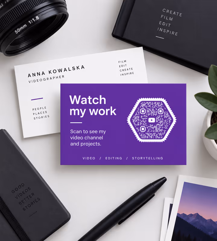 A purple card reading “Watch my work - scan to see my video channel and projects” with a white hexagonal QR code, on a desk with a camera lens and photo prints
