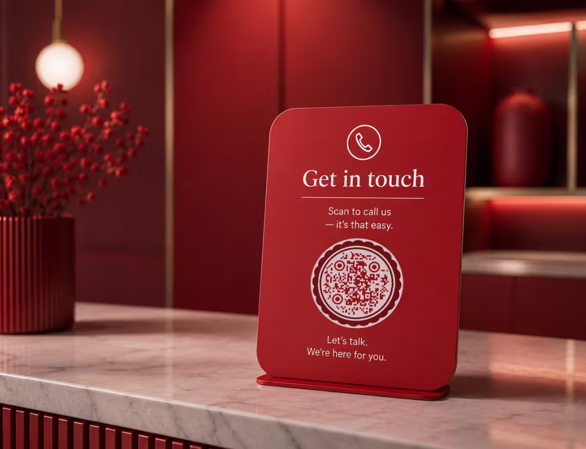 A red rounded “Get in Touch - scan to call us” table card with a circular QR code, on a marble counter beside a red vase