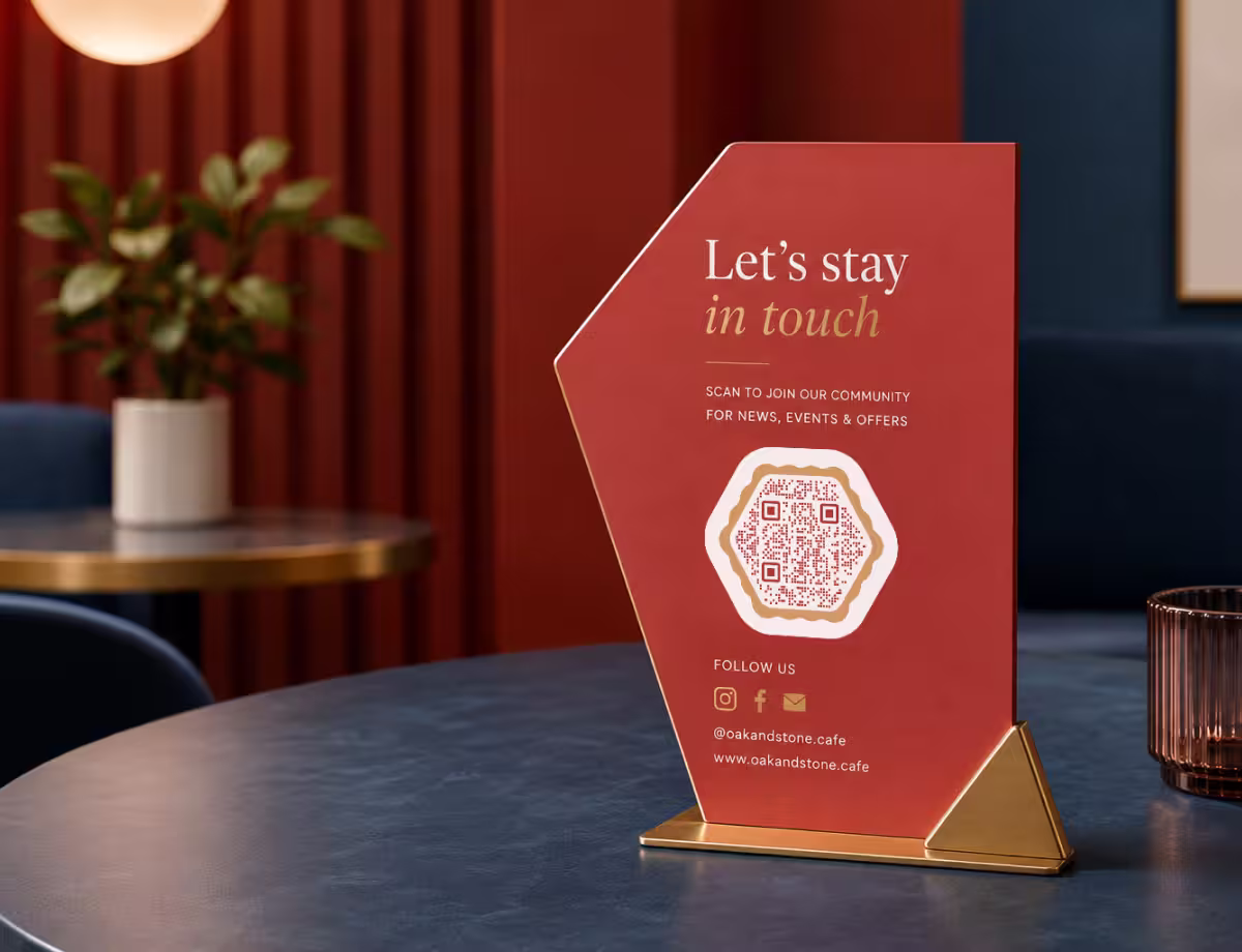 A terracotta hexagon-shaped “Let's stay in touch - scan to join our community” table display with a hexagonal QR code, on a brass stand