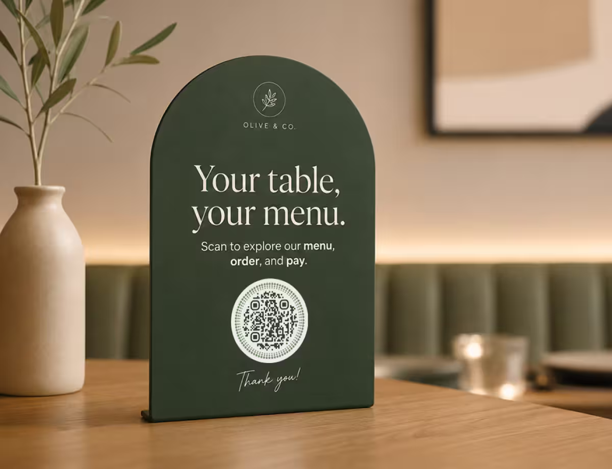 A dark-green arch-shaped “Your table, your menu - scan to order and pay” restaurant sign with a circular QR code, beside a cream vase