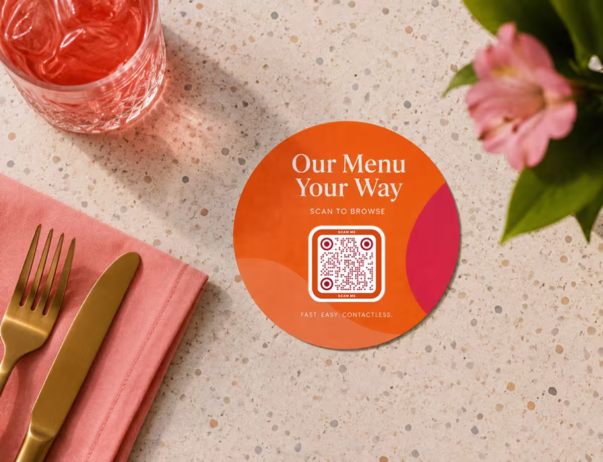 An orange round “Our Menu, Your Way - scan to browse” menu coaster with a QR code, on a table beside gold cutlery