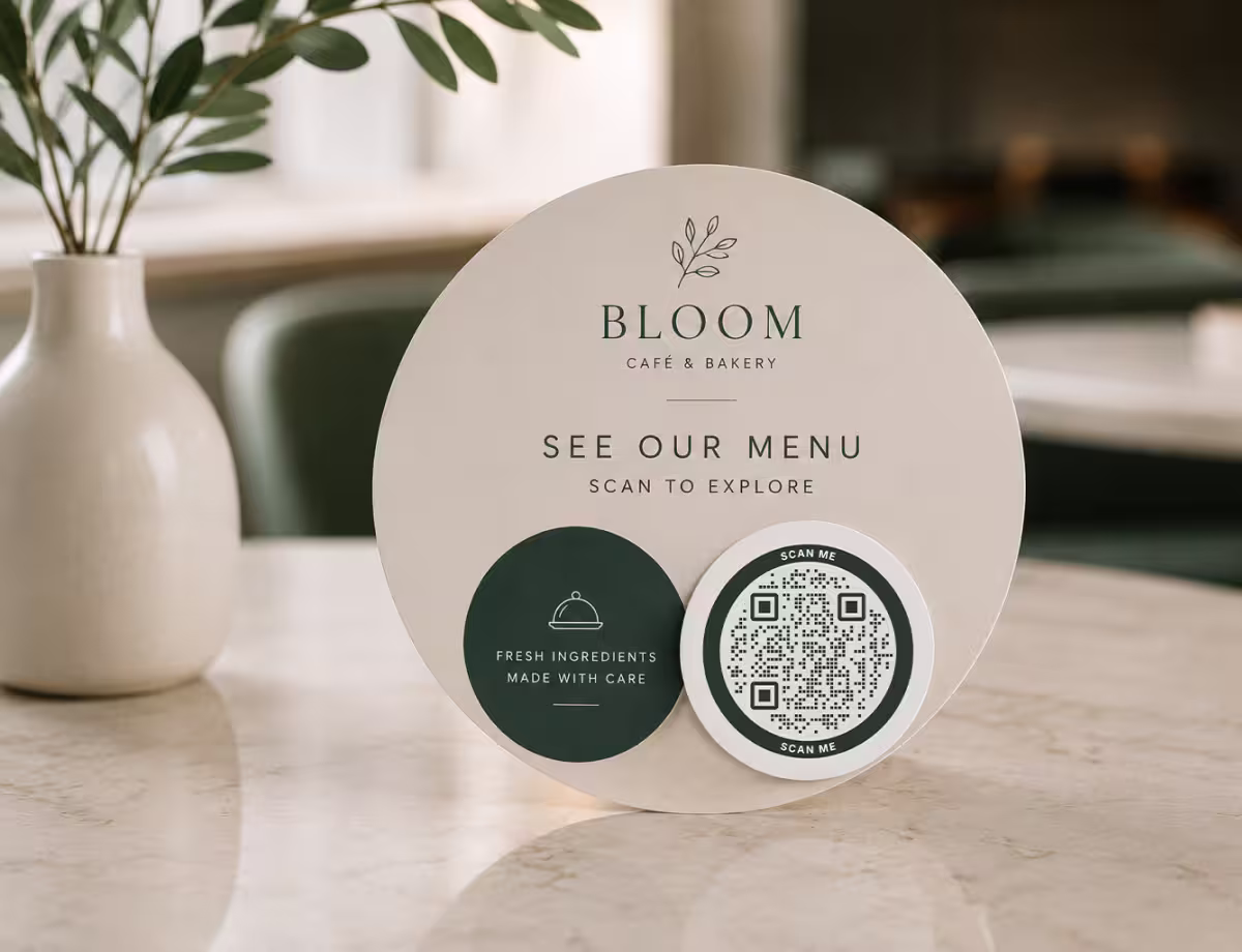 A round cream “Bloom Café - see our menu, scan to explore” table sign with a circular QR code, beside a white vase