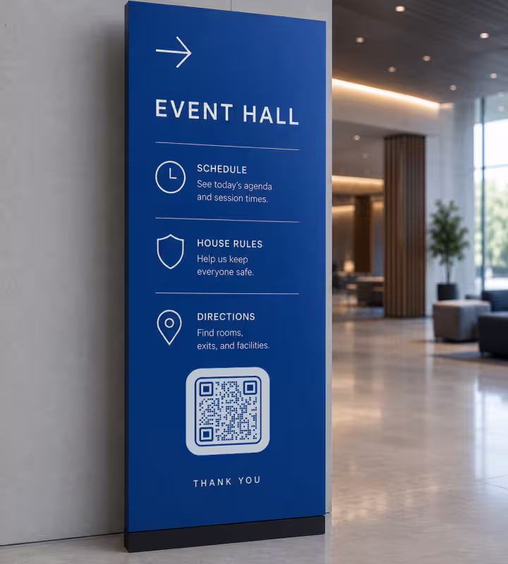 ป้ายบอกทางสีน้ำเงินทรงสูงข้อความ "Event Hall" แสดงกำหนดการและเส้นทางอยู่เหนือ QR Code ในล็อบบี้ของสถานที่จัดงาน