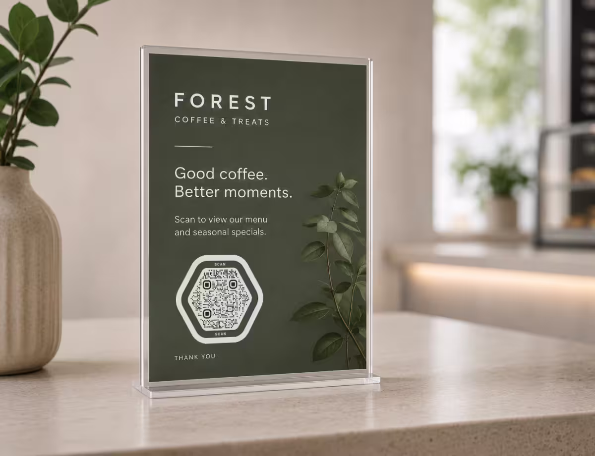 ป้ายตั้งโต๊ะอะคริลิกสีเขียวเข้ม "Forest Coffee & Treats - สแกนเพื่อดูเมนูของเรา" พร้อม QR Code หกเหลี่ยม ข้าง ๆ มีใบยูคาลิปตัสและแจกัน