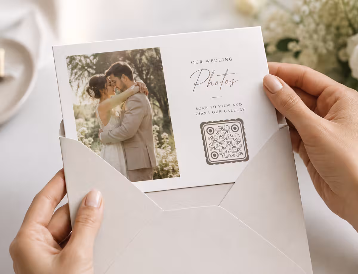 มือที่กำลังเปิดซองสีขาวซึ่งมีการ์ดรูปงานแต่งงานอยู่ข้างใน แสดงคู่บ่าวสาวกำลังโอบกอดกัน และ QR Code ทรงกลมที่มีข้อความ "สแกนเพื่อดูและแชร์แกลเลอรีของเรา"