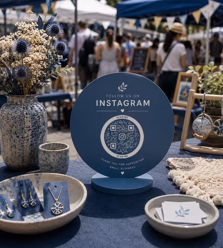 A round blue “Follow us on Instagram” sign with a QR code, on a craft-fair stall beside handmade jewellery