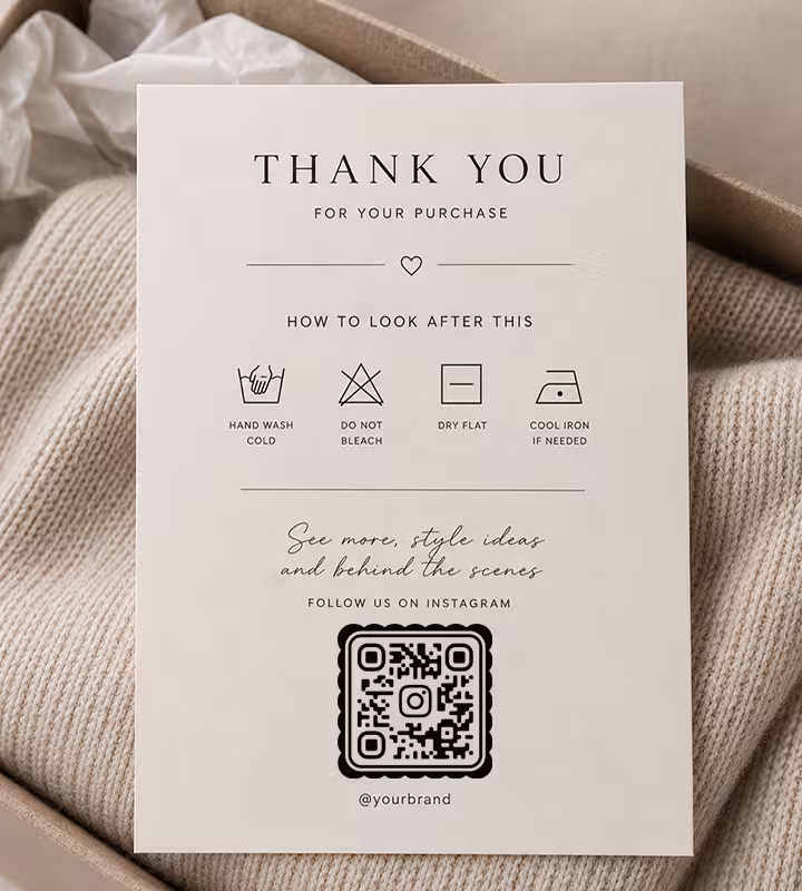 A cream “Thank you for your purchase” card with care symbols and a rounded QR code, resting on a folded knit in a gift box