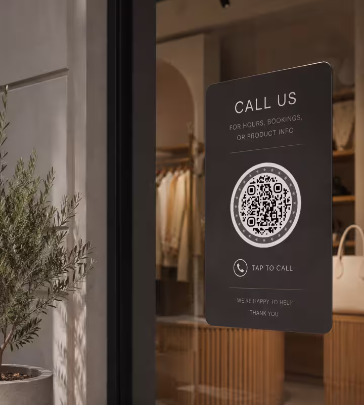 ป้ายสีเข้ม ข้อความ "โทรหาเรา - แตะเพื่อโทร" พร้อม QR Code บนประตูกระจกของร้าน มีต้นมะกอกอยู่ด้านหน้า