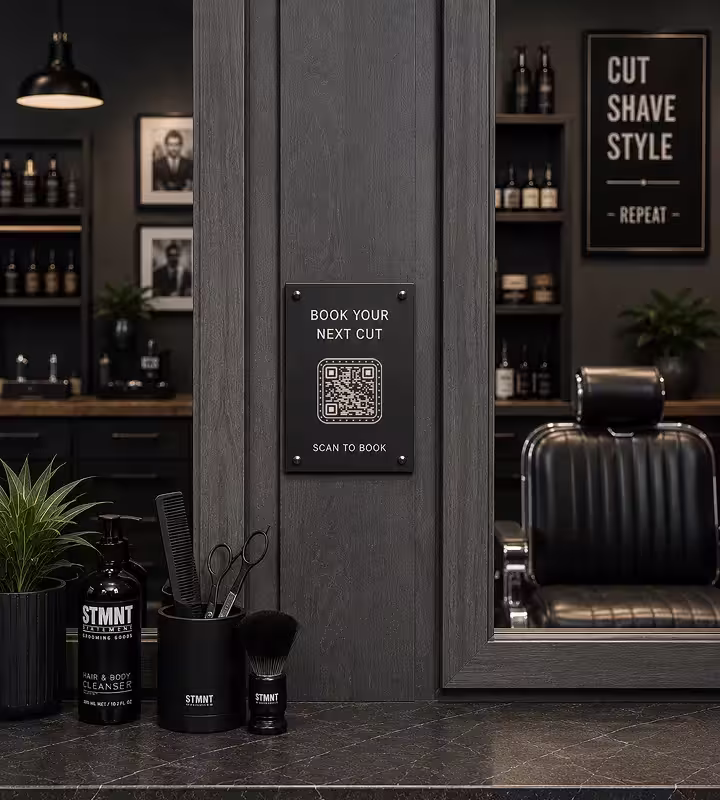 A small black plaque on the frame beside a barbershop mirror with a QR code reading “Book your next cut - scan to book”, above a counter of combs, brushes and grooming bottles