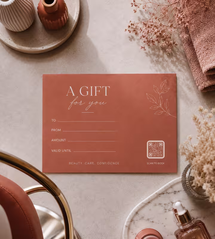 A terracotta gift voucher reading “A gift for you”, with blanks for the amount and expiry and a small QR code marked “Scan to book”, on a stone surface with a folded towel