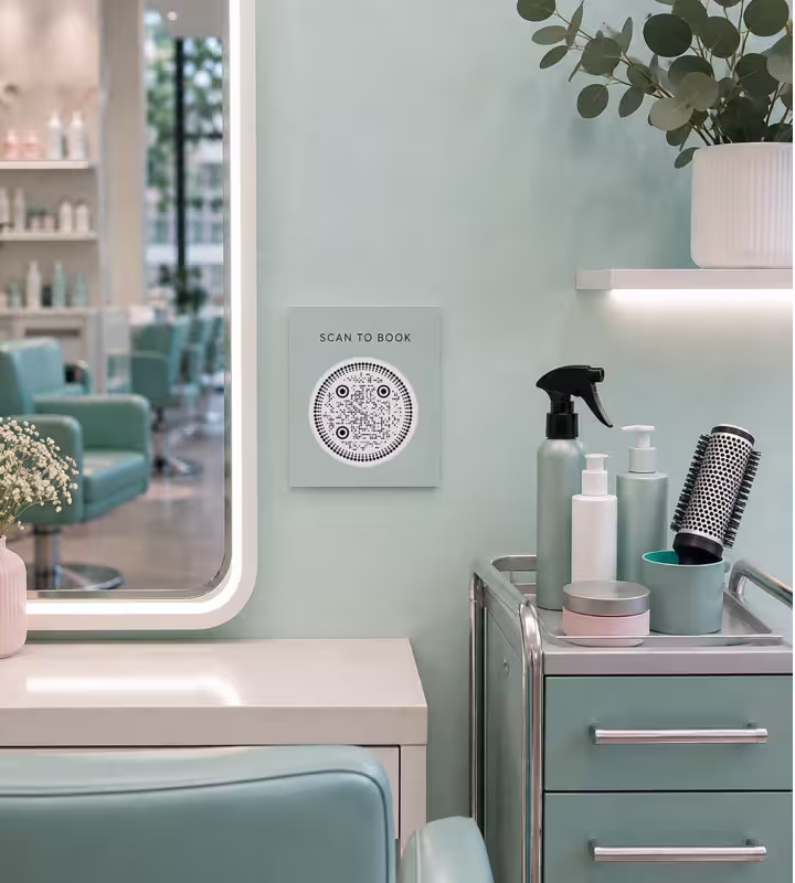 A small square sign on a mint-green salon wall reading “Scan to book”, with a round QR code, beside a lit mirror and a styling trolley of sprays and brushes