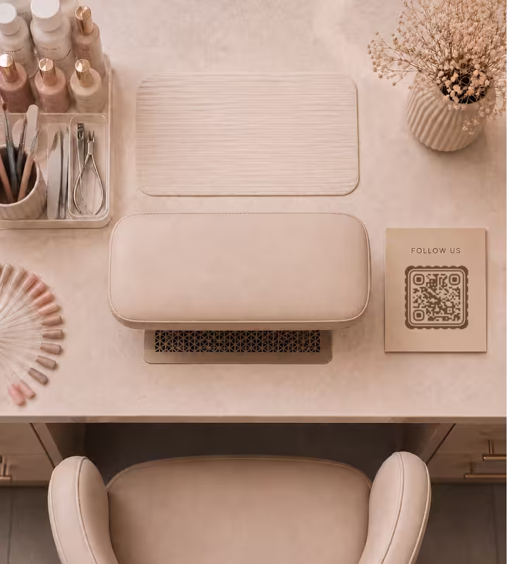 An overhead view of a beige nail desk with a “Follow us” card and a QR code beside the nail lamp, the tool tray and a fan of colour swatches