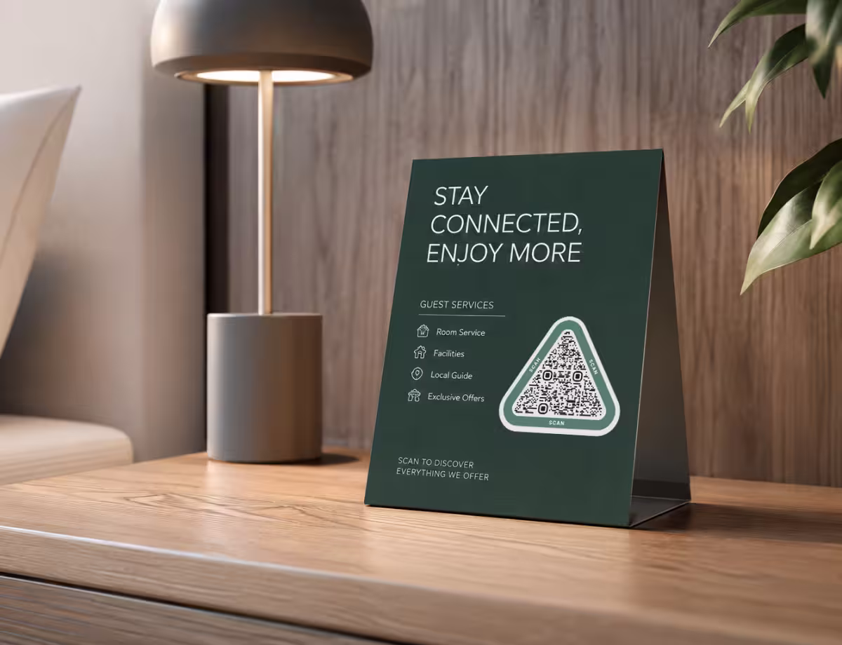 การ์ดข้างเตียงในโรงแรมสีเขียวเข้ม "Stay Connected, Enjoy More" ที่ระบุบริการสำหรับผู้เข้าพัก พร้อม QR Code ทรงสามเหลี่ยม วางบนโต๊ะข้างเตียงไม้ข้างโคมไฟ