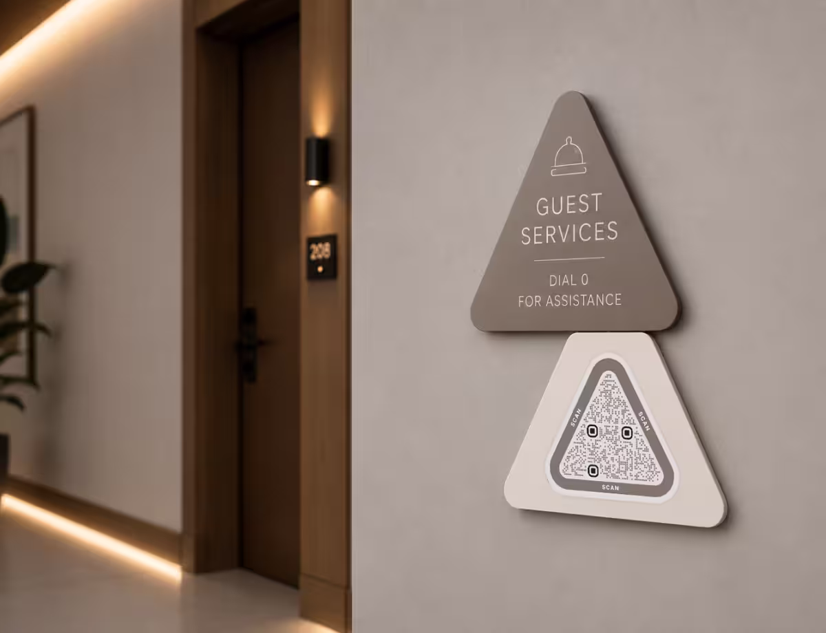 ป้ายผนังโรงแรมทรงสามเหลี่ยมสีเทา "Guest Services - กด 0 เพื่อขอความช่วยเหลือ" ติดอยู่เหนือ QR Code ทรงสามเหลี่ยม ข้างประตูห้องพัก