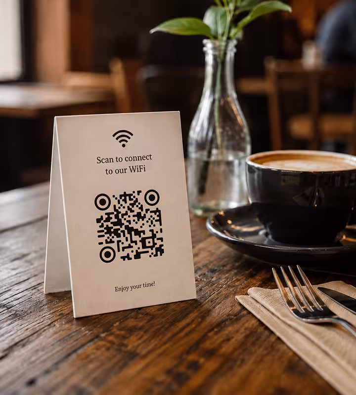 A folded table card on a wood café table reading “Scan to connect to our WiFi” with a QR code, beside a cup of coffee